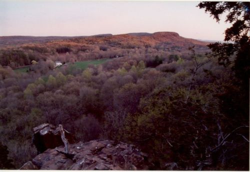 A scenic view of rolling hills and a valley, with autumn foliage and a distant mountain under a soft sunset sky.