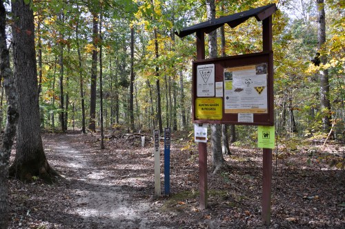 A wooded trail entrance with a signboard displaying information and rules, surrounded by autumn foliage.