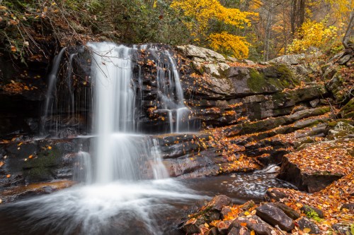 A serene waterfall cascades over rocky terrain, surrounded by autumn foliage and fallen leaves.
