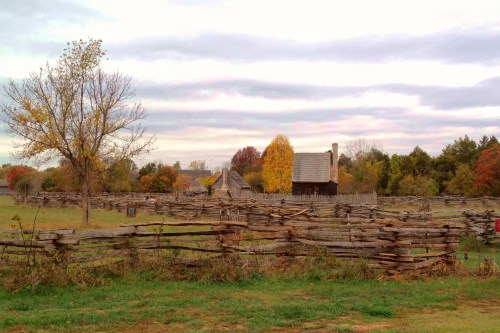 A rustic landscape featuring a wooden fence, autumn trees, and a historic building under a cloudy sky.
