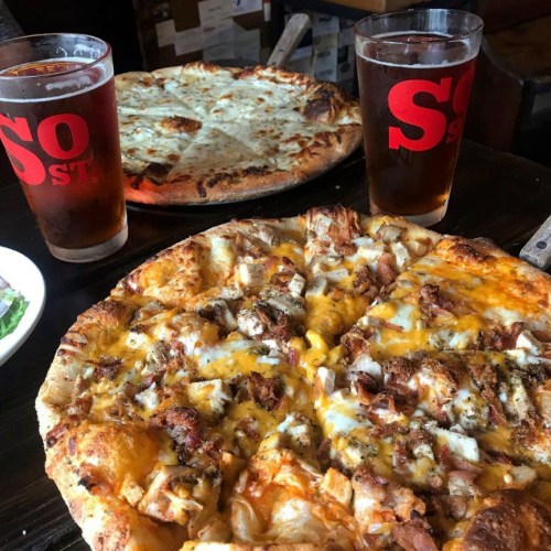 A close-up of a BBQ chicken pizza with two glasses of beer and a cheese pizza in the background.