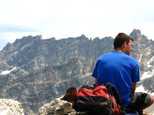A person sits on a rocky ledge, overlooking a mountainous landscape, with a backpack beside them.