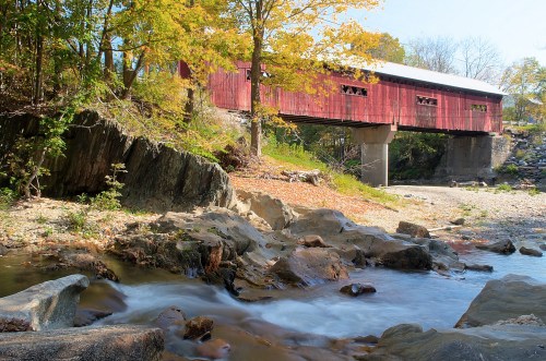 A red covered bridge spans a rocky stream, surrounded by autumn foliage and clear blue skies.