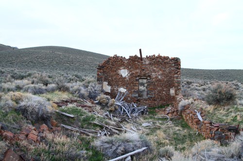 A dilapidated stone structure stands amidst overgrown grass and scattered debris, with rolling hills in the background.