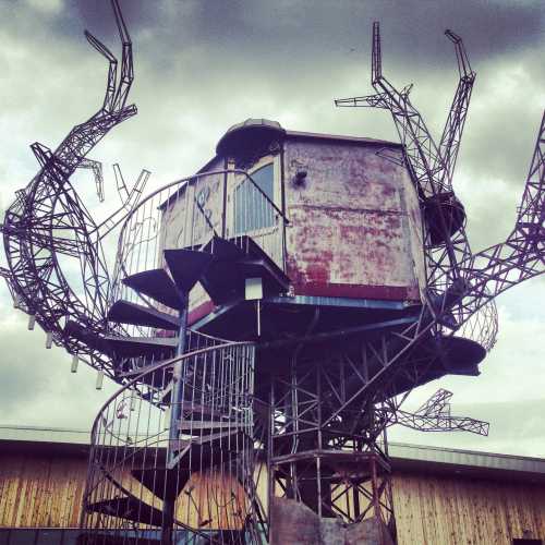 A large, metallic structure resembling a giant crab with a small building at its center, set against a cloudy sky.