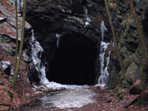 A dark cave entrance surrounded by icy stalactites and rocky terrain, with fallen leaves scattered on the ground.