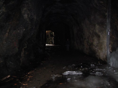 A dark, rocky cave with a faint light at the far end and ice scattered on the ground. A figure stands in the distance.