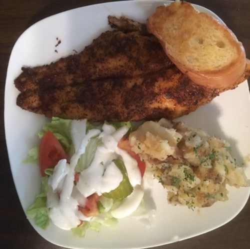 A plate with seasoned fish fillets, a slice of toasted bread, a side salad, and creamy potato salad.