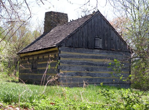 A rustic log cabin with a stone chimney, surrounded by greenery and trees in a peaceful outdoor setting.
