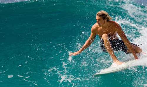 A shirtless surfer rides a wave, skillfully balancing on his board in vibrant blue water.