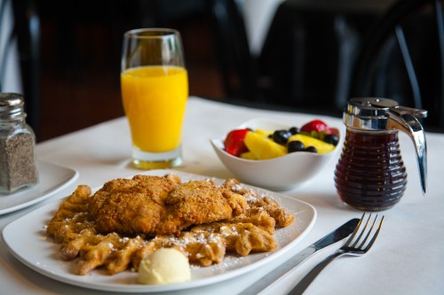 A plate of fried chicken and waffles, served with butter, syrup, a glass of orange juice, and a bowl of fresh fruit.