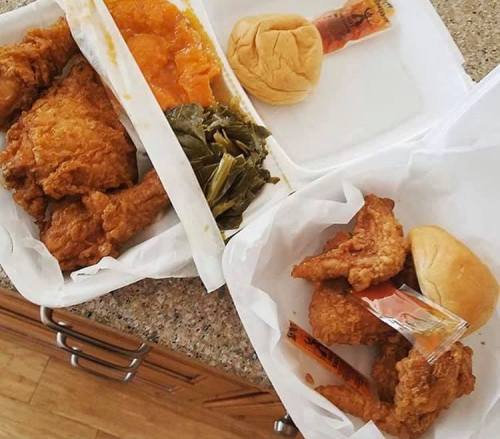 Two takeout containers with fried chicken, collard greens, a roll, and a side of orange sweet potatoes.