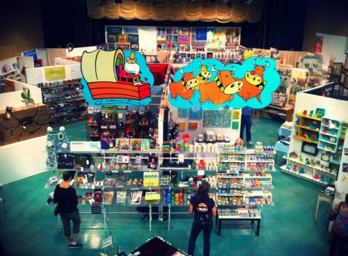A vibrant market scene with colorful displays, art, and animated characters above, showcasing various products and visitors.