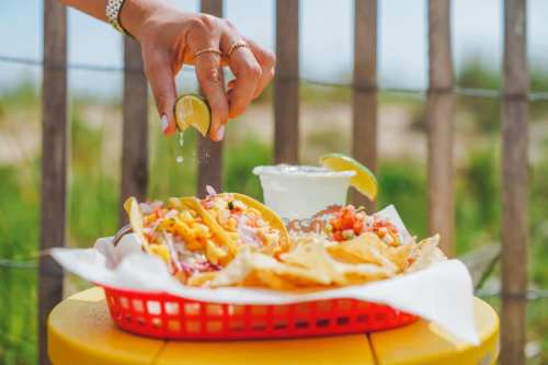 A hand squeezes lime over tacos and chips, with a margarita in the background, set against a sunny outdoor scene.