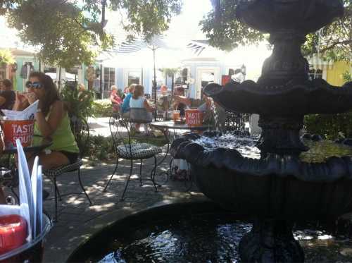 A sunny outdoor café scene with a fountain, tables, and people enjoying their meals in a vibrant setting.