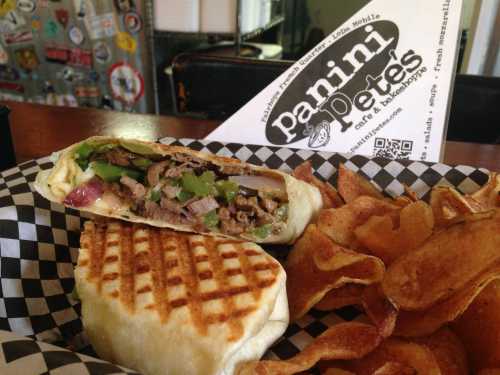 A grilled panini filled with meat and vegetables, served with crispy potato chips in a checkered basket.