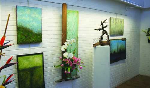 Art gallery display featuring abstract paintings, a sculpture, and floral arrangements against a white brick wall.