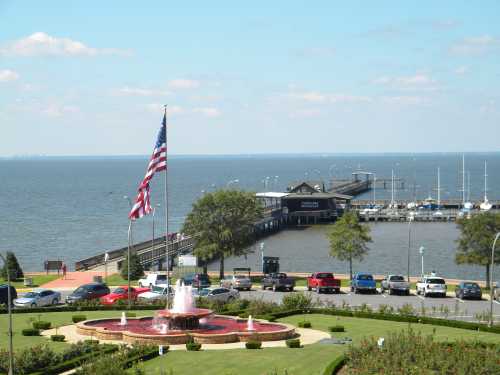 A waterfront view featuring a fountain, American flag, and a marina with boats, surrounded by green grass and parked cars.