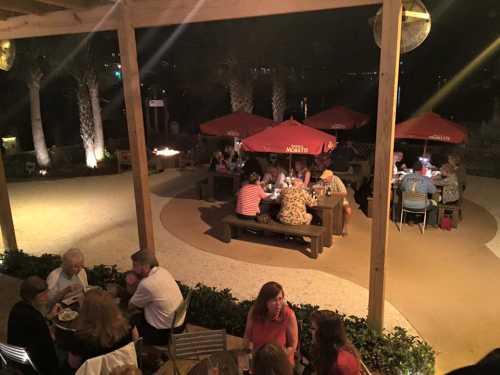 Outdoor dining area at night with people seated under red umbrellas, surrounded by palm trees and soft lighting.