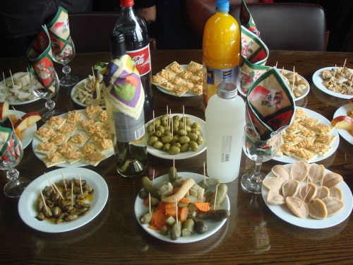 A table spread with various appetizers, drinks, and snacks, including olives, pickles, and crackers.