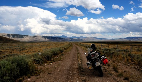 A motorcycle parked on a dirt road, surrounded by open fields and mountains under a cloudy sky.