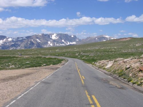 A winding road stretches through a grassy landscape, with mountains and blue skies in the background.