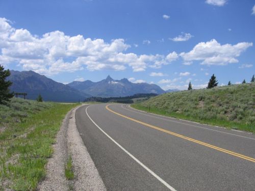 A winding road leads through green fields with mountains and a blue sky dotted with clouds in the background.