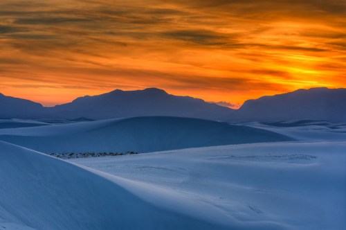 A stunning sunset over rolling white sand dunes, with mountains silhouetted against a colorful sky.