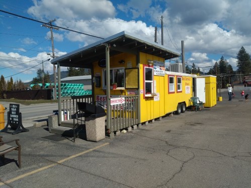 A bright yellow food stand with a porch, menu signs, and outdoor seating, set against a blue sky with clouds.