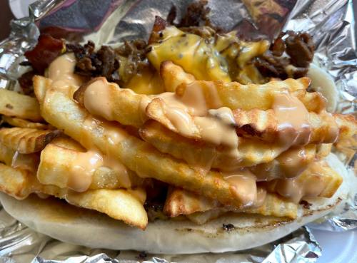 A loaded sandwich topped with crispy fries, melted cheese, and a drizzle of sauce, wrapped in foil.