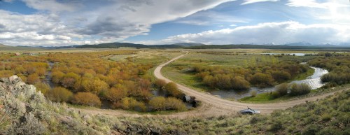 A panoramic view of a winding dirt road through a vibrant landscape of autumn foliage and a flowing river.