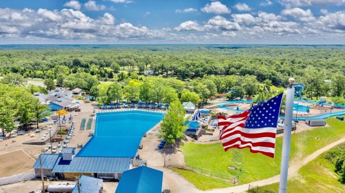 Aerial view of a water park with a large pool, slides, and a waving American flag against a blue sky and green trees.