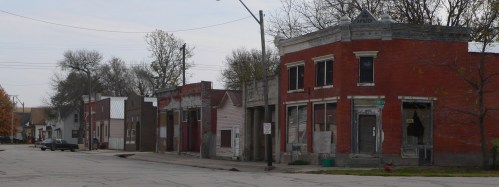 Abandoned buildings line a quiet street, showcasing weathered facades and overgrown trees in a small town.