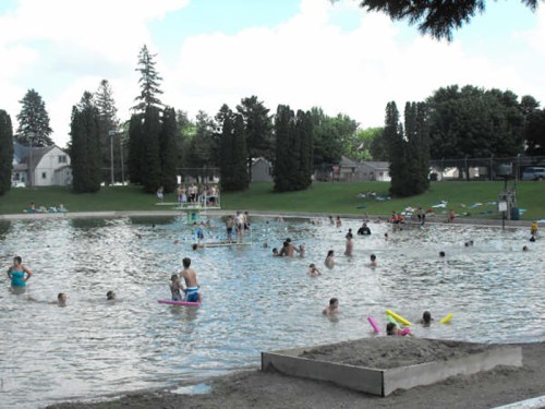 A crowded outdoor swimming area with people enjoying the water, surrounded by trees and grassy fields.