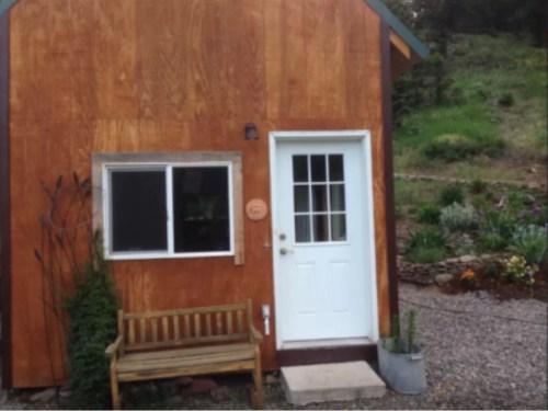 A small wooden cabin with a white door and window, surrounded by greenery and a gravel path. A bench is in front.