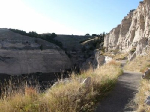 A narrow path winds through rocky cliffs and grassy terrain under a clear blue sky.