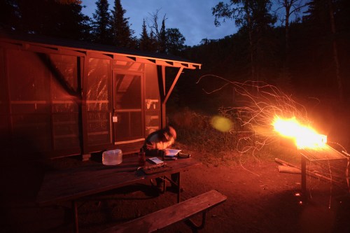 A campsite at dusk with a person near a table, sparks flying from a grill, and a cabin in the background.