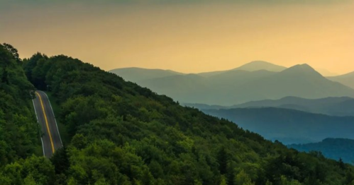 A winding road through lush green hills, with mountains fading into the distance under a soft sunset sky.