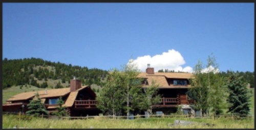 A large wooden house with a sloped roof, surrounded by trees and mountains under a clear blue sky.