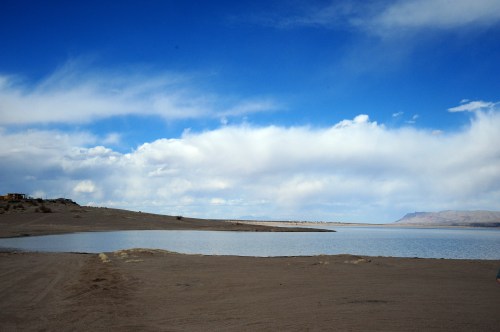 A serene landscape featuring a calm lake surrounded by sandy terrain and a blue sky with scattered clouds.