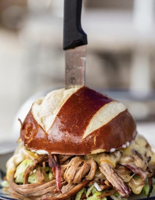 A large pretzel bun burger filled with pulled meat, cheese, and lettuce, topped with a knife.