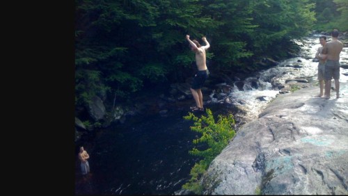 A person jumps off a rock into a river while two others watch nearby. Lush greenery surrounds the scene.