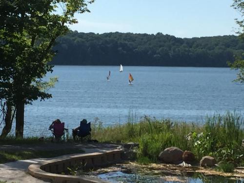 Two chairs by a lake with sailboats in the distance and lush greenery surrounding the water.