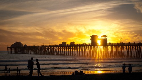 Silhouetted figures on a beach at sunset, with a pier and vibrant sky in the background.