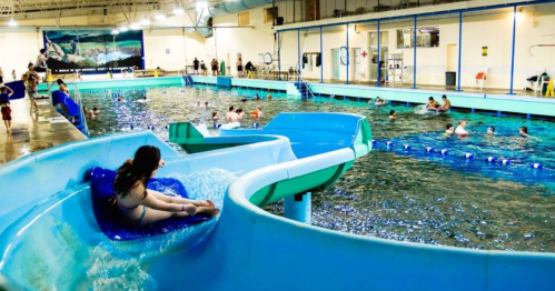 A water slide leads into a pool where people swim and relax in a brightly lit indoor aquatic center.