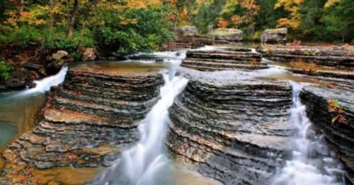 A serene waterfall cascades over layered rocks, surrounded by vibrant autumn foliage.