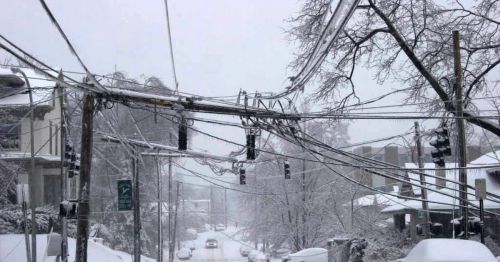 Snow-covered street with power lines and traffic signals, surrounded by trees and buildings in a winter landscape.