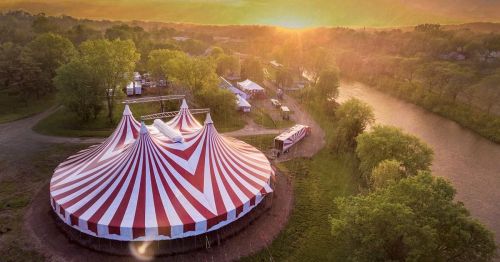 Aerial view of a striped circus tent at sunset, surrounded by trees and a river.