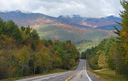 A winding road through vibrant autumn foliage, with mountains and cloudy skies in the background.