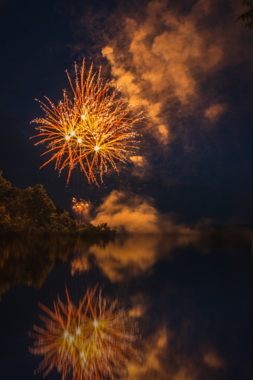Colorful fireworks burst in the night sky, reflecting on a calm water surface surrounded by trees.